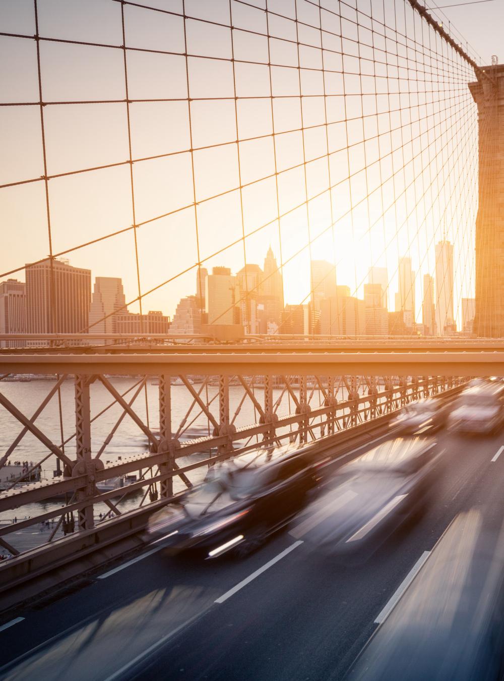 Brooklyn Bridge at sunrise, with cars whizzing across and people walking on the raised pedestrian walkway