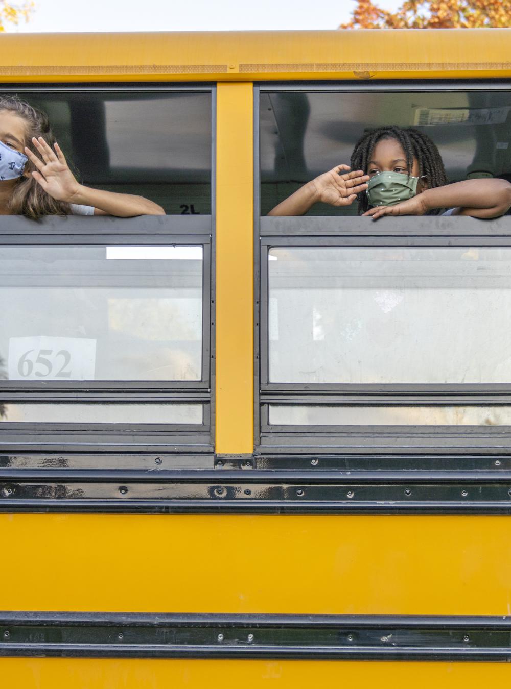 Two students in masks waving from an open school bus window