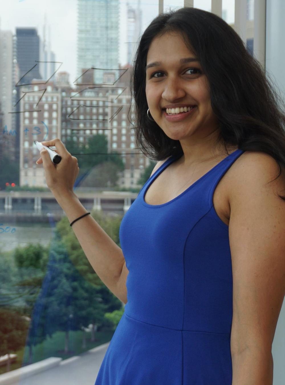 Shreya Chandrasekar writing on a window with a whiteboard marker, with a skyline behind her