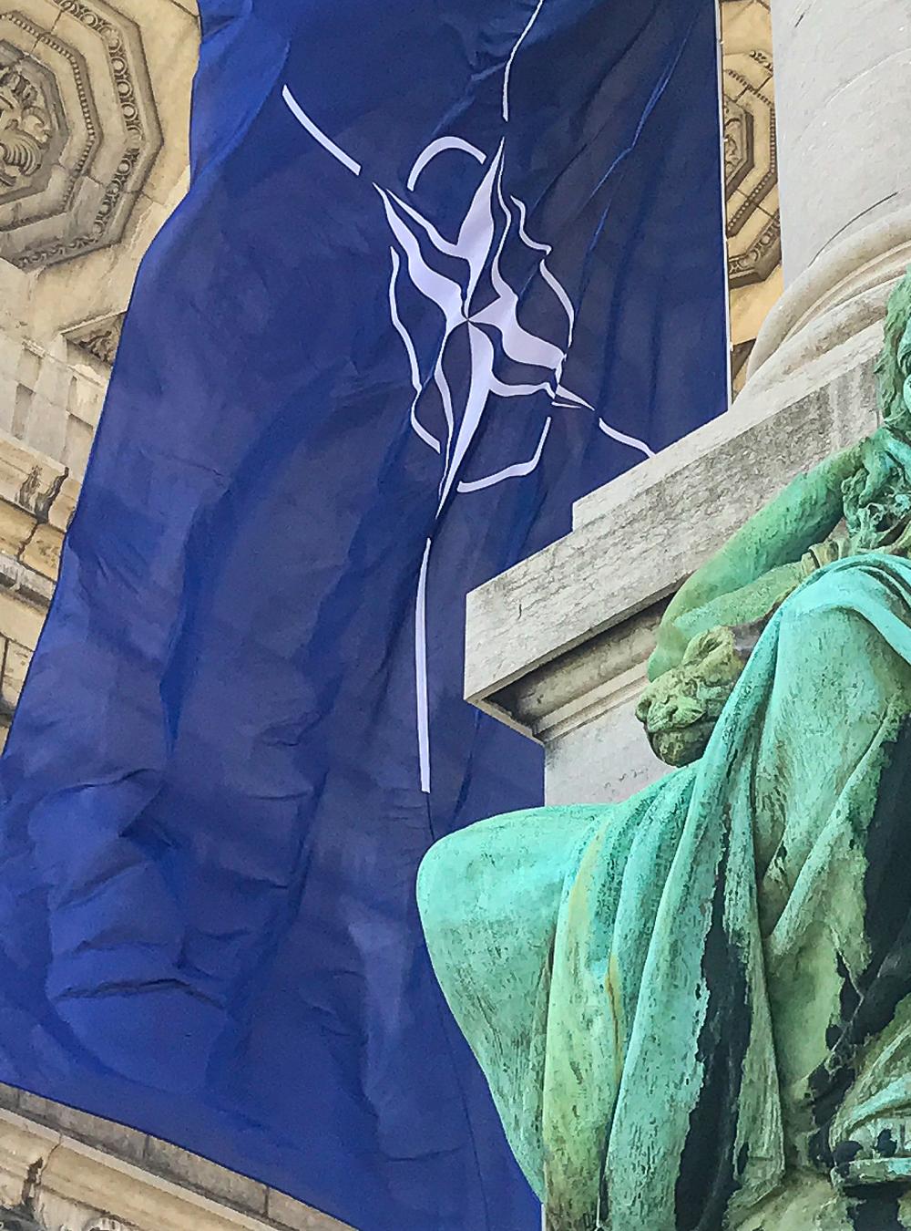The NATO flag flying under an arch with a statue in front
