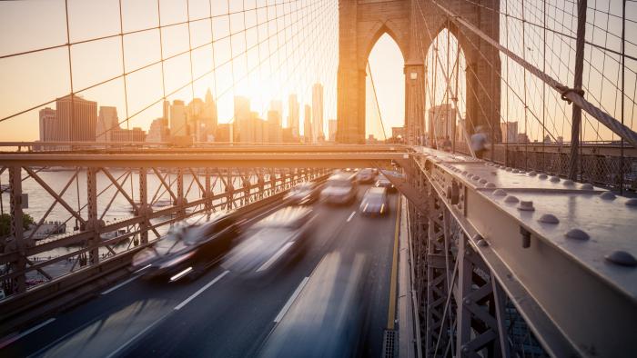 Brooklyn Bridge at sunrise, with cars whizzing across and people walking on the raised pedestrian walkway