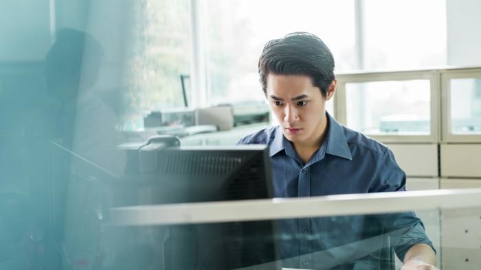 Viewed through glass, an employee with an invisible disability works at a computer in his office