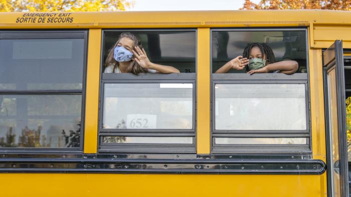 Two students in masks waving from an open school bus window