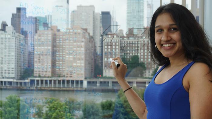 Shreya Chandrasekar writing on a window with a whiteboard marker, with a skyline behind her